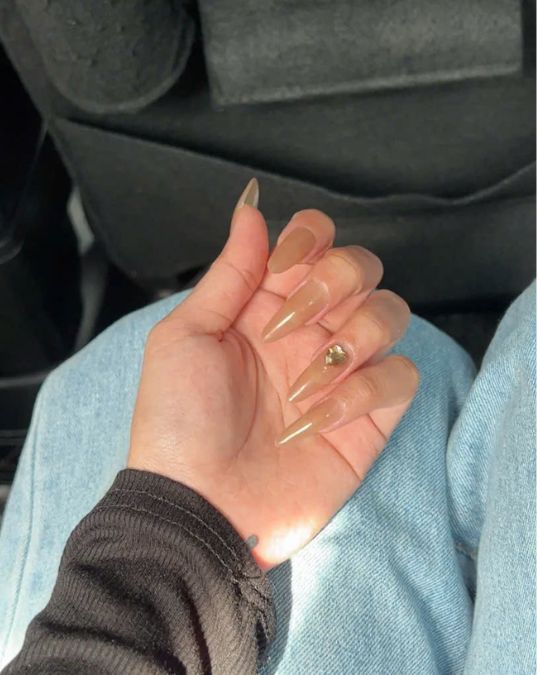 Long stiletto-shaped nails with nude beige gel polish and a single gold rhinestone accent on the ring finger, photographed against light blue denim and black fabric background in natural lighting
