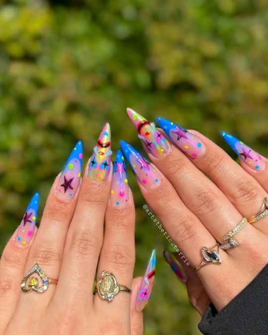Pointed stiletto nails with rainbow galaxy design, stars, and colorful dots.