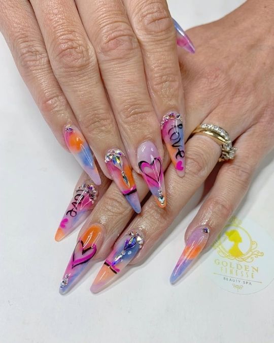 Vibrant sunset-inspired stiletto nails with love designs and crystal accents.